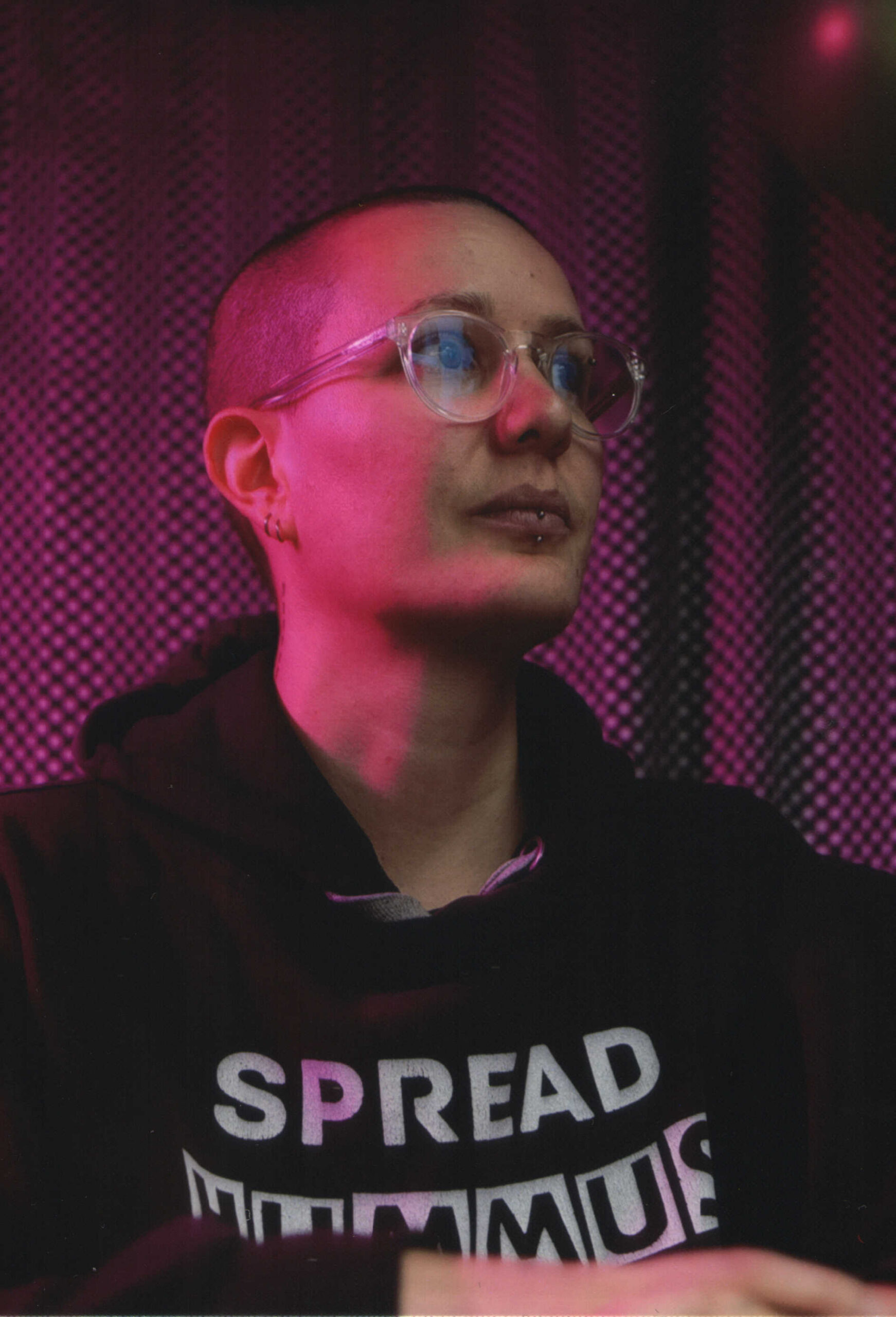Portrait de moi portant un sweat à capuche noir sur lequel il est écrit "Spread hummus not hate". J'ai le crâne rasé et des lunettes transparentes. La lumière est violette.